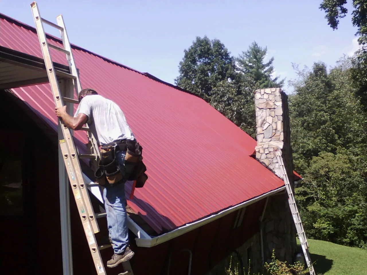 Roofer on a ladder beside a red metal roof and stone chimney — Soffit & Fascia Repair by Roofing Brothers in Fairburn